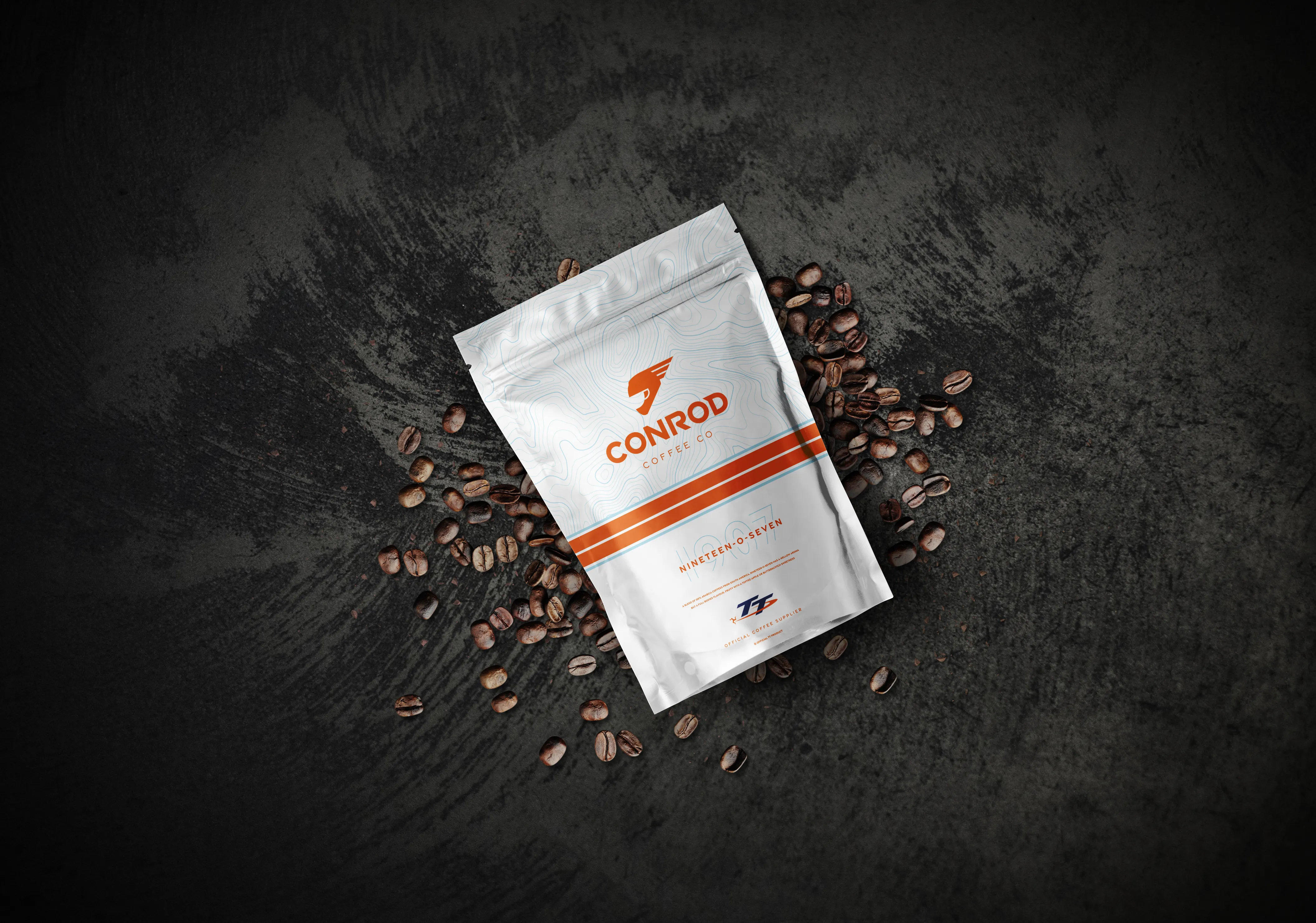 A white Conrod Coffee Co. bag with orange lettering is centered on a dark surface, surrounded by scattered roasted coffee beans, creating a rustic, aromatic feel.